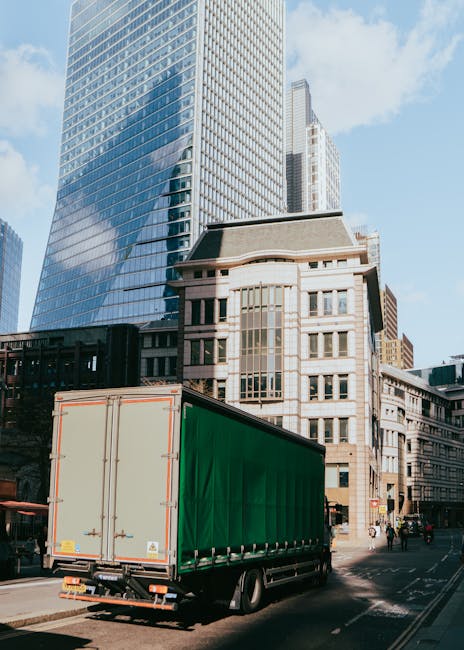 A large delivery truck with a green cargo container is parked on a city street amidst tall modern office buildings with glass facades and older architectural styles. The truck is positioned near a sid