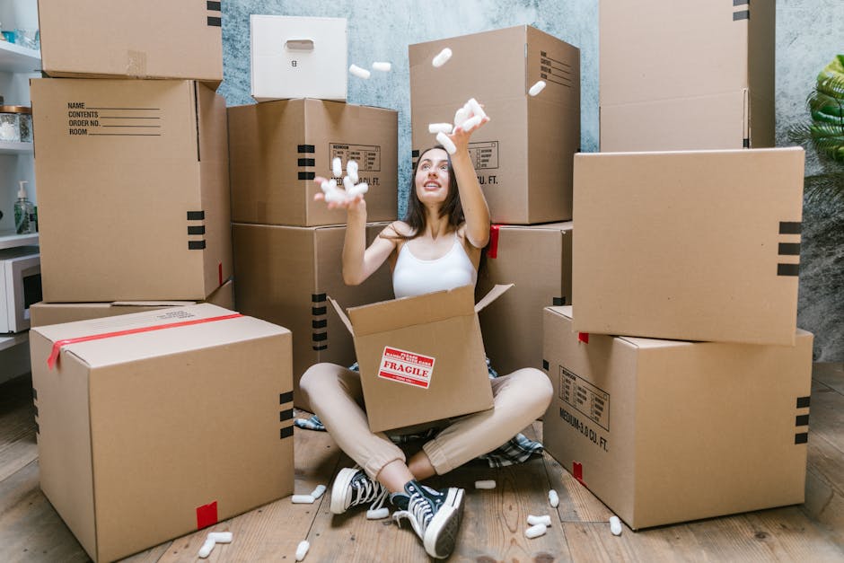 A young woman with long dark hair, wearing a white sleeveless top, beige pants, and black-and-white sneakers, sits on a wooden floor surrounded by numerous cardboard boxes of various sizes, some seale