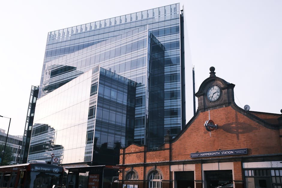 A modern glass office building with reflective windows towering above a historic brick station building with a clock and a sign reading 'Hammersmith Station,' situated on Uxbridge Road. In the foregro