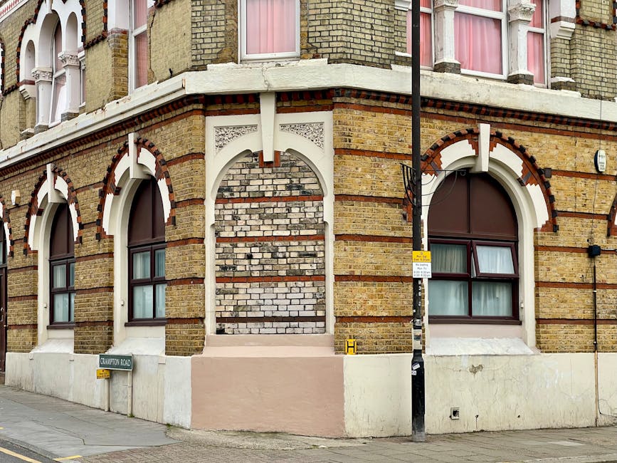 A modern glass office building with reflective windows towering above a historic brick station building with a clock and a sign reading 'Hammersmith Station,' situated on Uxbridge Road. In the foregro